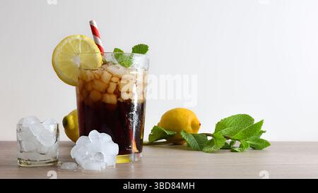 Erfrischendes Cola-Getränk mit Eis in einer Glasschale, dekoriert mit einem Strohhalm und einer Scheibe Obst auf einem Holztisch mit Obst, Eis und Minzblättern und einem Weiss Stockfoto