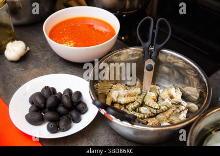Eine Metallschale roher Tigergarnelen mit Schere, schwarze Oliven und rote Tomatensauce, bereit für die Zubereitung von Meeresfrüchten. Stockfoto