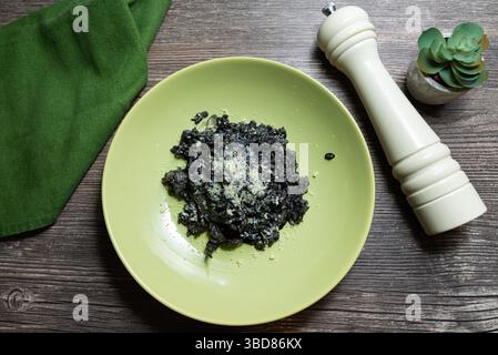 Blick von oben auf Tintenfisch-Risotto mit Parmesankäse, serviert in einer grünen Schüssel neben einer weißen Pfeffermühle auf Holztisch. Stockfoto