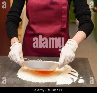 Person mit Handschuhen und Schürze, die während der Teigzubereitung eine Schüssel neigt, um Eigelb in einen Mehlhügel auf einer Küchentheke zu gießen. Stockfoto