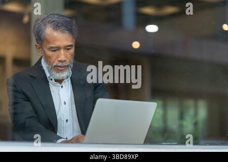 Älterer Mann, der sich auf Laptop in einem modernen Arbeitsbereich konzentriert Stockfoto