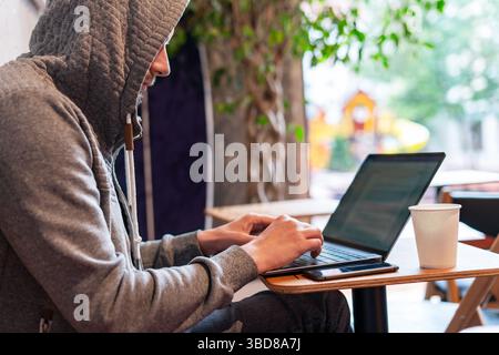 Webdesigner mit Kapuze sitzt am Kaffeetisch und arbeitet intensiv an einem Laptop. Die ruhige Umgebung lässt Konzentration vermuten, während die Kaffeetasse in der Nähe entspannt ist Stockfoto