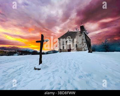 Die Schönheit einer schneebedeckten Landschaft mit einem Holzkreuz und einer malerischen Kirche, die in den warmen Farben des Sonnenuntergangs getaucht ist Stockfoto