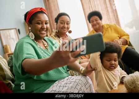 Gruppe von lächelnden Schwarzen mit Baby und Seniorin, die Selfie mit Smartphone machen, um sich im gemütlichen Wohnzimmer zu treffen Stockfoto