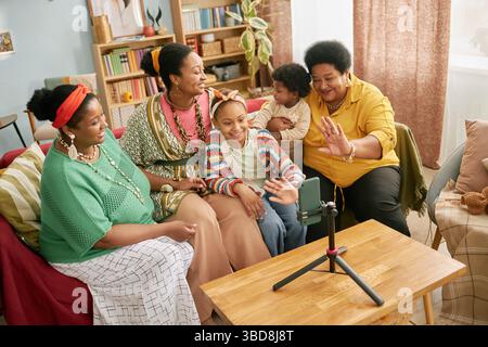 Die generationenübergreifende Black-Familie versammelte sich um Videoanrufe mit dem Telefon auf dem Stativ, lächelte, während sie sich lebhaft unterhalten und der Kamera im Wohnzimmer zuwinkte Stockfoto