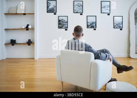 Mann sitzt im modernen Wohnzimmer und beobachtet gerahmte Kunstwerke an weißen Wänden. Bequem in gepolstertem Stuhl mit Holzfußboden Stockfoto