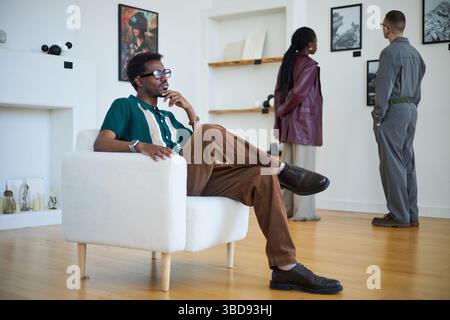Menschen, die zeitgenössische Kunstwerke in einer modernen Galerie untersuchen. Ein Mann saß in einem weißen Stuhl, während zwei Personen an der Wand die Stücke beobachten Stockfoto