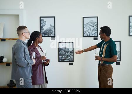 Gruppe von Erwachsenen, die über Fotos diskutieren, die in einer Kunstgalerie ausgestellt werden. Einzelpersonen halten Weingläser, während sie über das Kunstwerk sprechen, das an der Wa hängt Stockfoto