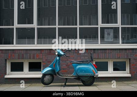 Vespa 50 Roller in Vintage-Blau parkt vor moderner Backstein- und Glasfassade in Hamburg. Stockfoto