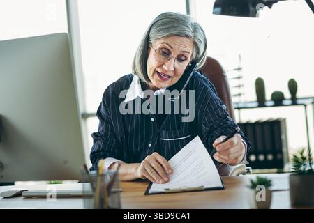 Selbstbewusste, erfahrene Geschäftsfrau multitasking in einer eleganten Büroumgebung und gleichzeitig effektive Verwaltung professioneller Dokumente Stockfoto