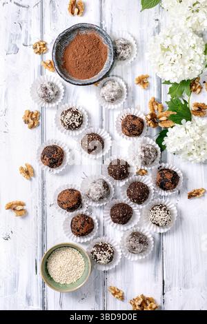 Hausgemachte Schokolade rohe vegane Kugeln Trüffel in Pappbecher Kakaopulver Sesamsamen Walnüsse und weiße Hortensie Blüten auf rustikalem weißem Holz Backgro Stockfoto