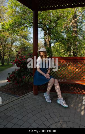 Ein Mädchen in blauem Kleid und weißer Mütze sitzt auf einer Holzbank in einem üppigen Garten, umgeben von Blumen und Bäumen, und genießt die friedliche Atmosphäre im Freien Stockfoto