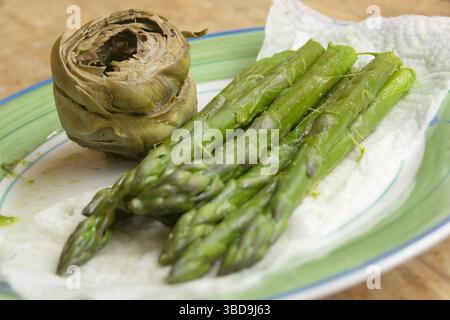 Aus nächster Nähe sehen Sie frische, leuchtend grüne Spargelstangen, die auf einem Papiertuch liegen, neben einem gekochten Artischockenkopf auf einem hellen Teller. Der Fokus Stockfoto