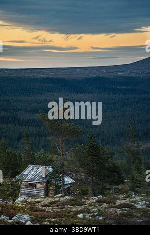 Eine verlassene Blockhütte, versteckt hinter Felsbrocken und einer Kiefer, während die Dämmerung den orange-blauen Himmel über den dichten Wald unten in Dalarna Schweden wirft Stockfoto