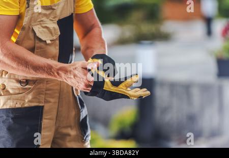 Ein Bauarbeiter zieht Sicherheitshandschuhe an und bereitet sich auf eine Aufgabe in einem lebhaften Außenbereich mit viel Grün und Werkzeug vor. Der Mitarbeiter ist konzentriert Stockfoto
