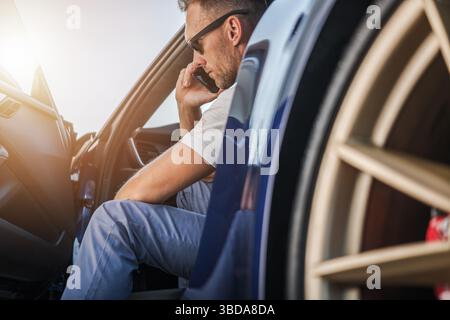 Ein lässig gekleideter Mann sitzt auf dem Fahrersitz eines Autos und führt ein Telefongespräch, während die goldenen Sonnenstrahlen die Szene erleuchten. Stockfoto
