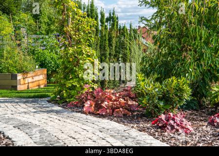 Ein gewundener Steinweg führt durch einen üppigen Garten voller bunter Laub und lebhafter grüner Pflanzen. Die Szene wird durch helles Sonnenlicht beleuchtet Stockfoto
