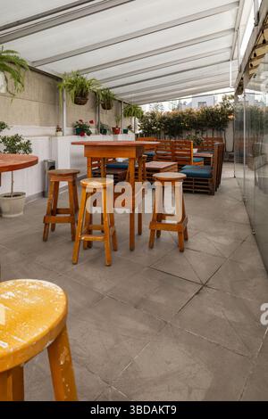 Eine gemütliche Außenterrasse mit Holztischen, Stühlen und Barhockern unter einem Baldachin bietet einen zwanglosen und einladenden Raum für gesellschaftliche Veranstaltungen. Stockfoto