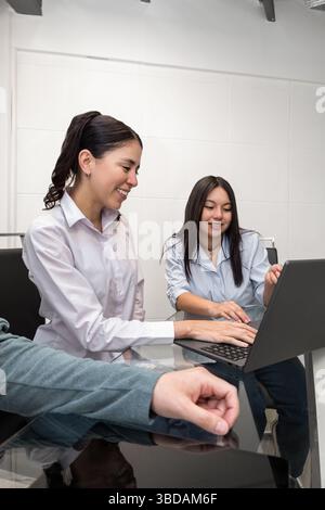 Zwei junge Frauen sitzen zusammen an einem Tisch, lächeln, während sie auf einen Laptop schauen und reflektieren Teamarbeit, Freundschaft und positive Energie in einer modernen Umgebung. Stockfoto