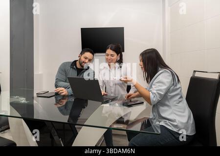 Drei junge Frauen sitzen um einen Tisch, konzentrieren sich auf ein Notebook und führen eine lebhafte Diskussion, die Teamarbeit, Zusammenarbeit und kreatives Brainsto symbolisiert Stockfoto