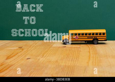 Ein gelber Spielzeugschulbus steht auf einem Holztisch vor einer grünen Tafel mit dem Satz „Back to School“ in weißen Buchstaben, represen Stockfoto