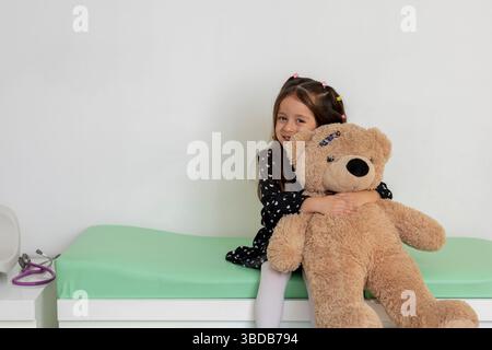 Glückliches lateinisches Mädchen, das ihren Teddybären umarmt, während er auf einem medizinischen Untersuchungstisch im Kinderarztbüro sitzt Stockfoto