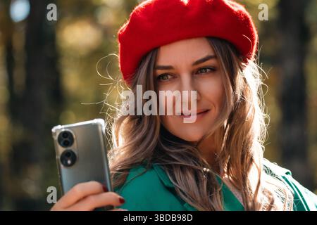 Frau Selfie Smartphone Herbst draußen: Rote Baskenmütze, lächelnde Frau macht Selfie mit Smartphone im Herbst. Stockfoto