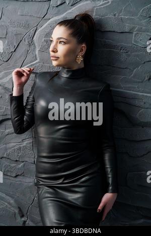 Eine auffällige junge Frau mit olivfarbenem Teint, trägt ein elegantes schwarzes Rollkragenkleid aus Leder und goldene Ohrringe und posiert selbstbewusst gegen einen Text Stockfoto