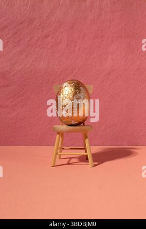 Goldenes Ei auf Holzstuhl mit rosafarbenem Hintergrund. Minimalistische Komposition Stockfoto