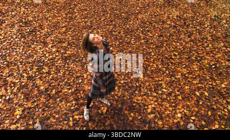 Luftaufnahme einer attraktiven jungen Frau, die auf dem Waldboden aus Blättern in Herbstfarben steht. Erhöhtes Foto des Modells. Stockfoto