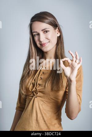Die junge schöne Frau zeigt das OK-Zeichen auf dem grauen Hintergrund Stockfoto
