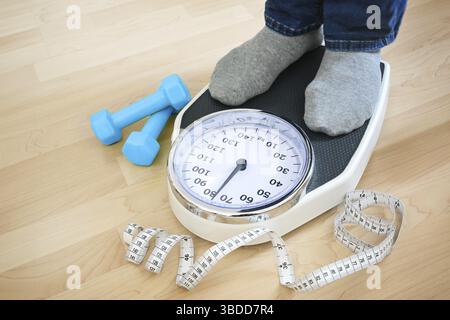 Füße eines Mannes in grauen Socken, die auf einer Waage stehen, um das Gewicht nach dem Fitnesstraining zu überprüfen, blaue Kurzhanteln und ein Maßband, das in der Nähe auf einem Wald liegt Stockfoto