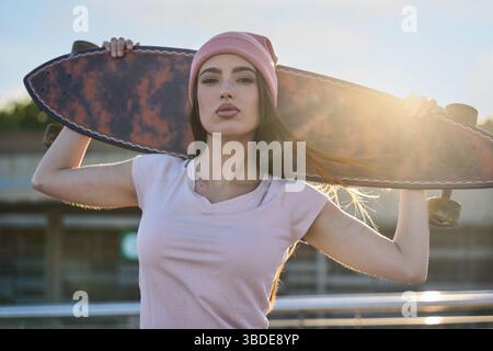 Stilvolle Frau, die bei Sonnenuntergang Longboard hält und den urbanen Lebensstil annimmt Stockfoto