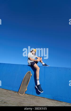 Junger Skater, der auf einer blauen Wand mit seinem Skateboard unter klarem Himmel ruht Stockfoto