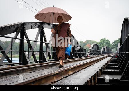 Buddhistischer Mönch in braunen Gewändern trägt einen rosa Regenschirm, während er über die historische Brücke über den Fluss Kwai in Kanchanaburi, Thailand, spaziert. Stockfoto