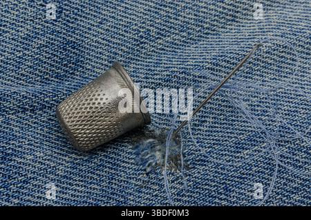 Nahaufnahme einer Metallkausche, einer Nähnadel mit Garn und einem Denim-Stoff mit sichtbaren Näharbeiten, die traditionelle Textil-Repai symbolisieren Stockfoto