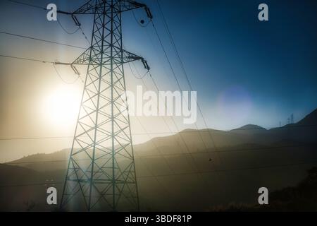 Dramatisches Bild eines Kraftübertragungsturms, der sich vor einem sonnendurchfluteten Nebelhimmel befindet, mit teilweise in Dunst gehüllten Hügeln im Hintergrund Stockfoto