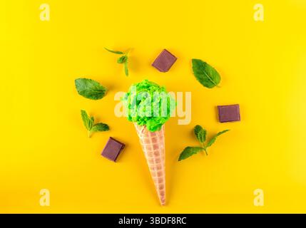 Ein grüner Eiskegel mit Minzblättern und Schokoladenscheiben ist auf einem gelben Hintergrund platziert. Der Kegel ist umgeben von Minzblättern und Schokoladen-waf Stockfoto
