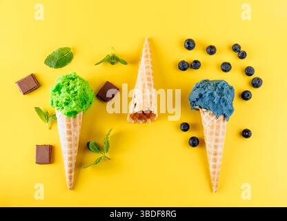 Eine Tischansicht von drei Eiskegeln mit verschiedenen Geschmacksrichtungen auf gelbem Hintergrund. Die Kegel sind mit Heidelbeeren, Schokoladenstückchen und verziert Stockfoto