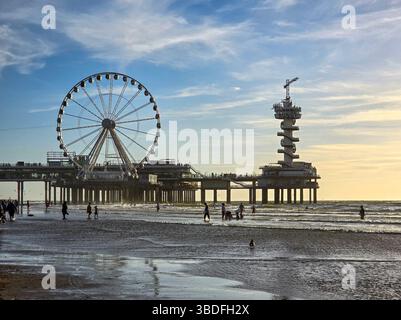 Die Menschen schlendern entlang eines Sandstrandes in der Nähe eines Piers mit einem großen Riesenrad und Turm, das in den sanften Glanz eines farbenfrohen Sonnenuntergangs an der Küste getaucht ist Stockfoto