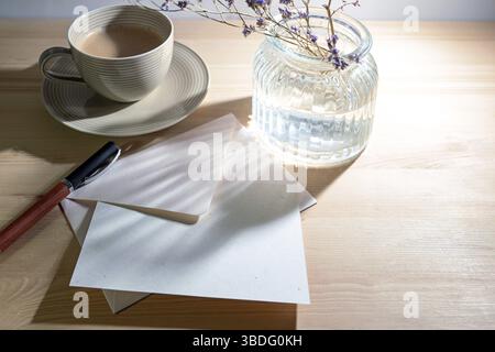 Grußkarte und Umschlag aus natürlichem Recyclingpapier, Stift, Kaffeetasse und eine Glasvase mit Sonnenlichtreflexionen am Morgen auf einem Holztisch, W Stockfoto
