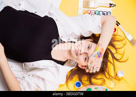 Fröhliche Künstlerin liegt auf gelbem Hintergrund, von Hand bedeckt mit Farbe, umgeben von Pinseln und Palette. Studio Pause, Kunst Lifestyle, Stockfoto