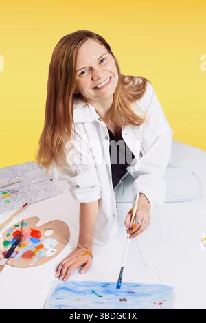 oman malt auf dem Boden mit einer Palette und einem Pinsel, lächelt vor Freude vor einem kräftigen gelben Hintergrund, kreativer Lebensstil, Maltherapie Stockfoto