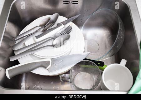 Teller, Löffel, Messer, eine Tasse und eine Schüssel stapeln sich nach Gebrauch in einem Metallwaschbecken und warten darauf, gewaschen zu werden, zu spülen, zu Haushaltsarbeiten Stockfoto