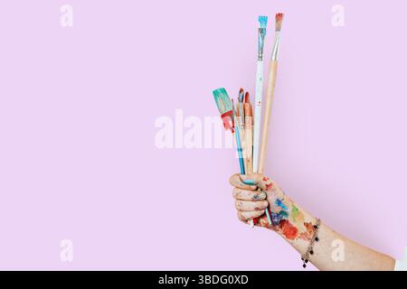 Eine Künstlerhand, die mit Farbe bedeckt ist und verschiedene bunte Pinsel vor einem kräftigen rosa Hintergrund hält, künstlerische Werkzeuge, kreativer Prozess Stockfoto