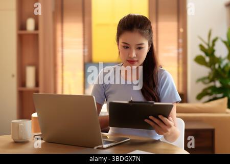 Eine junge asiatische Frau arbeitet von zu Hause aus mit einem Laptop und hält ein Klemmbrett. Sie konzentriert sich auf ihre Aufgaben, während sie an einem Holztisch sitzt und an einem gemütlichen geldautomaten sitzt Stockfoto