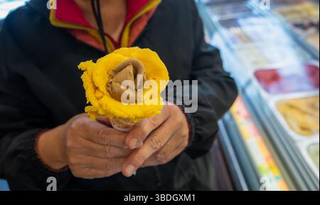 Eine Hand hält einen wunderschön gefertigten rosenförmigen Eiskegel mit gelben Mango- und braunen Schokoladenaromen und einer Vitrine mit verschiedenen eissorten Stockfoto
