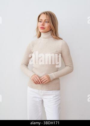 Eine junge Frau mit langen blonden Haaren, die einen beigen gerippten Rollkragenpullover und eine weiße Hose trägt und nachdenklich zur Seite blickt. Ihre Hände sind sanft Stockfoto