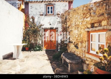 Sonniger Innenhof in Aldeia da Mata Pequena, Portugal, mit einem rustikalen weiß getünchten Haus mit einer lebhaften orangen Holztür, Steinmauern und Kletterpflanze Stockfoto