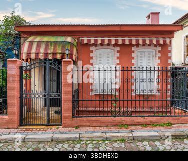 Außenansicht eines kleinen orangen Hauses mit weißen Fensterläden, schwarzem Metallzaun und gestreiften Markisen auf einer kopfsteingepflasterten Straße, tagsüber Stockfoto